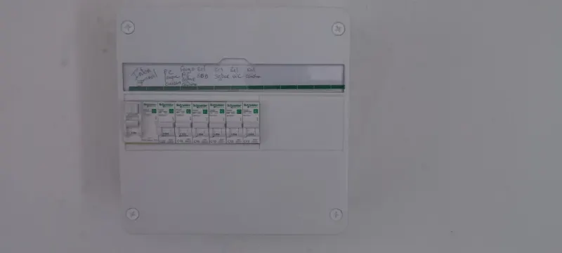 Un petit tableau électrique, chaque disjoncteur annoté à la main : plaque de cuisson, frigo, éclairage de la salle de bain, du séjour, des WC, de la cuisine.